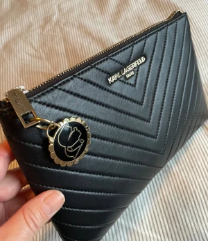 Svart quiltad kuvertväska Karl Lagerfeld - Stilren svart kuvertväska från Karl Lagerfeld med quiltad design i V-mönster och guldfärgad logga framtill. Väskan har dragkedja med dekorativt hänge och ett rymligt fack med mönstrat innerfoder. Perfekt för att bära med det viktigaste. (Bara lite sliten inuti runt dragkedjan.)