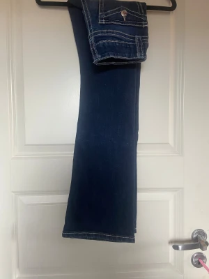 Gina Tricot Mörkblå bootcut jeans med kontrastsömmar - Säljer ett par mörkblå jeans med bootcut passform och tydliga vita kontrastsömmar. Jeansen har fickor med lock och knappdetalj bak, klassisk femficksdesign och sitter snyggt i midjan. Materialet är jeans och de har en normal passning.