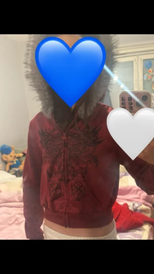 2000th vintage kofta  - Säljer en röd hoodiejacka från Carpe Diem med grå fuskpäls runt luvan. Jackan har ett stort svart tryck med vingar och text på framsidan, dragkedja och croppad passform. Perfekt för dig som gillar edgy streetstyle och detaljer.