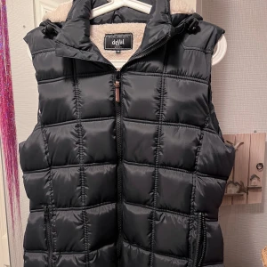 Svart quiltad dunväst från deVal - Svart quiltad dunväst från deVal med dragkedja framtill, huva och mysigt beige foder. Västen har två fickor med dragkedja och en snygg detalj med logopatch nertill. Perfekt för lager-på-lager och chill höst- och vinterstil.