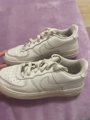 Nike Air Force 1 vita sneakers 36.5 (23.5cm) - Säljer ett par klassiska Nike Air Force 1 sneakers i helvitt skinn med rund tå och platt sula. De har perforerade detaljer på tån, snörning och den ikoniska swoosh-loggan på sidorna. Perfekta för dig som gillar clean och tidlös streetstil.