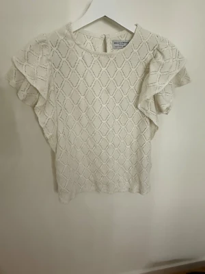 Vit blus med volang från Holly & Whyte (by Lindex) - Supersöt vit blus från Holly & Whyte by Lindex i storlek S. Blusen har ett snyggt rutmönster, korta volangärmar och rund hals. Materialet är hyfsat mjukt och bekvämt, perfekt för vår och sommar. Stängs med knapp i nacken. Perfekt att styla med jeans eller kjol!