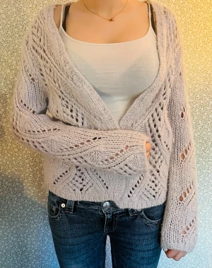 Ljusgrå stickad kofta med mönster - Supermysig ljusbeige stickad kofta med öppet hålmönster och breda ribbade muddar. Koftan har en loose passform och långa ärmar, perfekt att slänga över en topp för extra lager. Snyggt och chill plagg för höst och vinter.
