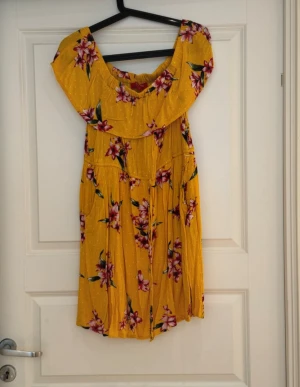 Gul blommig kortklänning från Guess - Superfin gul kortklänning från Guess med blommigt mönster i rosa och grönt. Klänningen har offshoulder-krage med volang och är tillverkad i mjuk bomull. Perfekt för sommarens alla tillfällen, och den har en luftig och skön passform.