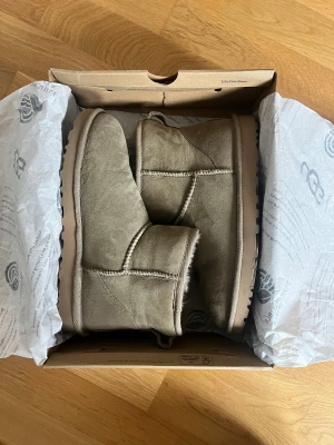 Beige UGG boots i mocka - Säljer ett par klassiska UGG boots i beige mocka med rund tå och platt sula. Insidan är fodrad med mjuk päls för extra värme och komfort. Skorna har synliga sömmar och UGG-logga baktill. Perfekta för kalla dagar och har en chill vibe.