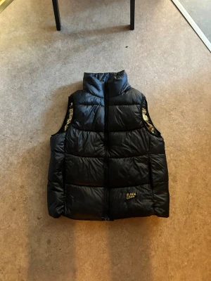 Svart dunväst från Black Good - Svart dunväst från Black Good med gulddetaljer och quiltad design. Västen har hög krage, dragkedja framtill och insida i glansigt guldtyg med hexagonmönster. Perfekt för lager-på-lager och ger en cool streetstyle-vibe.