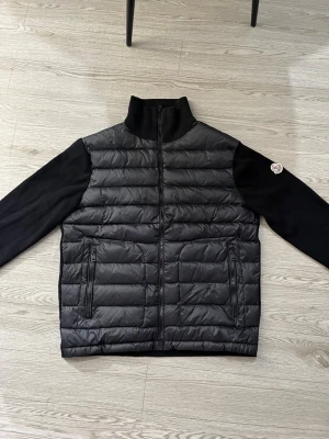 Svart Moncler pufferjacka med stickade ärmar - Svart pufferjacka från Moncler med stickade ärmar och hög krage. Jackan har quiltad front i syntetmaterial, två dragkedjeförsedda fickor och Moncler-logga på vänster armen denna jacka passar bra inför vår/höst den har en stilren och sportig