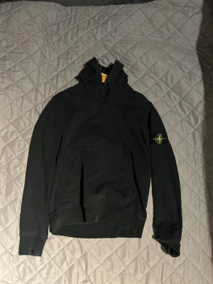 Svart hoodie från Stone Island - Svart hoodie från Stone Island med klassisk patch på vänster ärm. Tröjan har huva, magficka och ribbade muddar. Materialet är mjuk bomull och passformen är normal. Perfekt för dig som gillar streetwear och vill ha en clean look.