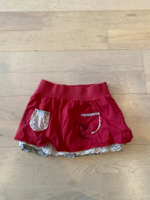 Röd kortkjol Dancing Queen KappAhl - Röd kortkjol från KappAhl med bred resår i midjan, två fickor fram med blommigt och hundtandsmönstrat tyg, samt texten 'Dancing Queen' tryckt på framsidan. Kjolen har volangkant i blommigt tyg nedtill för en extra söt look.