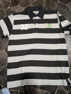 Randig pikétröja Färjestad BK XL - Snygg pikétröja från Färjestad BK i svart och grått med breda ränder. Tröjan har klassisk krage, knappar framtill och broderat klubbmärke på bröstet. Tryckt Färjestad-text på ärmen ger extra sportig vibe. Perfekt för dig som gillar hockey och vill visa ditt lag .Ny endast provade