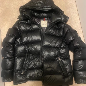 Svart Moncler pufferjacka med huva - Moncler Dunjacka – Storlek 3 (M/L)  Säljer en klassisk svart Moncler-dunjacka i storlek 3. Jackan är varm, lätt och har den typiska glansiga finishen som Moncler är kända för. Modellen har avtagbar huva, två snedställda fickor med dragkedja och rejäl tvåvägsdragkedja framtill.  Skick: Jackan är i bra användbart skick, men har några mindre skador/slitage, vilket syns på de sista bilderna: 	•	Ett litet hål nära nederdelen av dragkedjan 	•	Slitage/uppskavning vid en söm i samma område  