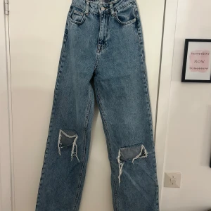 Blå ripped jeans från NA-KD, wide fit - Säljer ett par blå jeans från NA-KD med hög midja och wide fit. Jeansen har slitna hål vid knäna för en cool, avslappnad look. Klassisk femficksmodell och tillverkade i jeansmaterial. Perfekta för dig som gillar trendiga och bekväma byxor.
