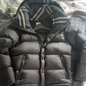 Burberry jacka  - Snygg svart dunjacka från Burberry med klassiskt rutigt foder i huvan. Jackan har dragkedja och knappar framtill, två fickor med dragkedja och en patch pả ärmen. Perfekt för kalla dagar och riktigt stilren look.