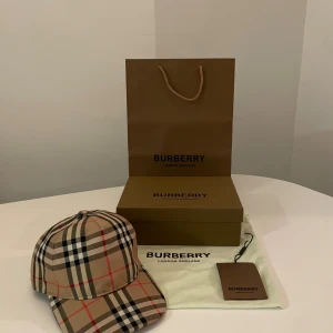 Burberry rutig keps i beige och svart - Snygg keps från Burberry med klassiskt rutigt mönster i beige, svart, vitt och röda detaljer. Justerbar baktill med svart spänne och Burberry London England-logga. Tillverkad i bomull, perfekt för dig som vill ha en ikonisk streetstyle-accessoar.