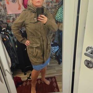 Olivgrön parkajacka från Ellos  - Säljer en olivgrön parkajacka från Ellos med huva, stora fickor och dragsko i midjan. Strl 38/40 .  Jackan har långa ärmar och stängs med dragkedja och knappar. Perfekt för dig som vill ha en snygg och praktisk jacka till vardags.