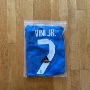 Blå Real Madrid Vini Jr. tröja Adidas S - Snygg blå Real Madrid fotbollströja från Adidas med Vini Jr. och nummer 7 på ryggen. Tröjan har vita detaljer, korta ärmar och klassiska Adidas-ränder på axlarna. Materialet är lätt och andas, perfekt för match eller träning. Emirates Fly Better tryck på bröstet.