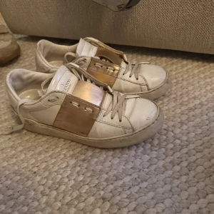 Valentino Garavani sneakers med guld - Snygga vita sneakers från Valentino Garavani med bred metallic guldpanel över snörningen. Klassisk låg modell med rund tå, platt sula och diskreta nitar bak. Skorna är i skinn och har vita snören. Perfekta för dig som vill sticka ut med lyxig detalj.