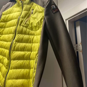 Gulgrön pufferjacka från Parajumpers - Snygg pufferjacka från Parajumpers i limegrönt och olivgrönt med huva och dragkedja. Jackan har quiltad framsida, släta ärmar och en bröstficka med dragkedja. Märkespatch på ena ärmen ger extra attityd. Perfekt för dig som vill sticka ut. Storleken är y-m vilket motsvarar 14 år 