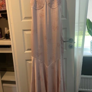 Ljusrosa långklänning med blommor - Säljer en ljusrosa långklänning med subtila blommönster och. Klänningen har en mjuk, glansig yta och en snygg söm under bysten som ger en feminin siluett. Perfekt för dig som gillar romantiska och eleganta vibes.