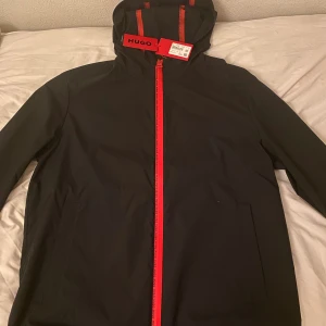 Svart vindjacka från Hugo Boss med röd dragkedja - Svart vindjacka från Hugo Boss med röd dragkedja och röda detaljer. Jackan har huva, två sidofickor och en innerficka med röd Hugo-logga. Snygg och stilren design som sticker ut med de röda accenterna. Perfekt för dig som gillar streetwear med premiumkänsla.