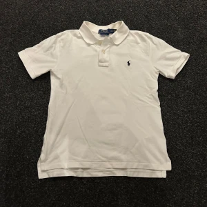 Vit pikétröja Ralph Lauren - Klassisk vit pikétröja från Ralph Lauren med broderad logga på bröstet. Tröjan har korta ärmar, krage och knäppning framtill. Tillverkad i mjuk bomull och har en tidlös, clean look som passar perfekt till jeans eller shorts.