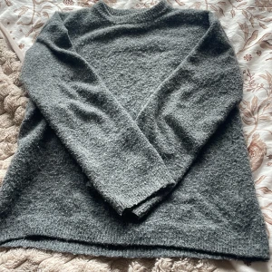 Mörkgrå stickad tröja från H&M - Mysig mörkgrå stickad tröja från H&M med rund halsringning och långa ärmar. Tröjan har en fluffig och lite ruggad yta som ger en avslappnad vibe. Perfekt för kyliga dagar och enkel att matcha med jeans eller kjol.