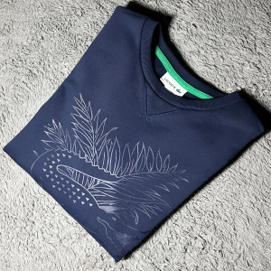 Mörkblå Lacoste tröja Roland Garros  - Mörkblå tröja från Lacoste Roland Garros-kollektionen med stor krokodil och palmblad tryck framtill. Klassisk rund hals, ribbade muddar och broderad Lacoste-logga vid nederkanten. Skön bomullsblandning och långärmad modell.