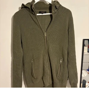 Olivgrön cardigan  - Olivgrön stickad  cardigan   med hdragkedja framtill och två fickor med dragkedja. Huva i quiltad polyester ger en cool kontrast till den stickade looken. Perfekt för lager-på-lager under hösten och vintern.