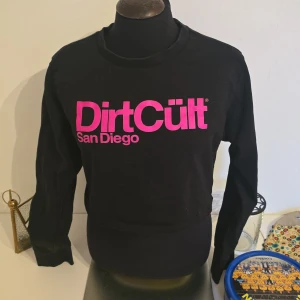 Svart sweatshirt DirtCült S - Svart sweatshirt från DirtCült med fet rosa logga 'DirtCült San Diego' framtill. Tröjan har rund hals, långa ärmar. Lite kortare modell