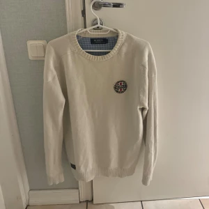 Vit stickad tröja från Morris - Vit stickad tröja från Morris med rund halsringning och broderad logga på bröstet. Klassisk och stilren design med ribbade muddar vid ärmslut och nederkant. Perfekt för lager-på-lager under kyliga dagar.