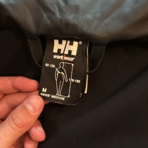 Svart regnjacka från Helly Hansen - Svart regnjacka från Helly Hansen Workwear i storlek medium. Jackan har huva, hel dragkedja framtill och två stora fickor. Materialet är syntetiskt och vattenavvisande, perfekt för blöta dagar. Enkel och stilren design med HH-logga på nedre delen.