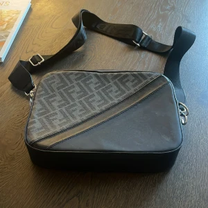 Svart Axelväska Från Fendi i Skinn - Säljer en svart axelväska från Fendi med bred justerbar rem och klassiskt Fendi-mönster på ena sidan. Väskan är tillverkad i skinn och har detaljer i silverfärgad metall samt dragkedja. Perfekt för dig som vill ha en stilren och lyxig accessoar. Den är lite “sliten” på baksidan men inget som syns när man har den på sig.