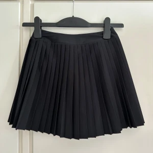 Svart plisserad kortkjol från Stadium - Svart plisserad kortkjol från Stadium med klassisk tennisstil. Kjolen har bred linning och är sydd i ett stretchigt material av polyester och elastan för en bekväm passform. Perfekt för dig som gillar sportig och stilren look.