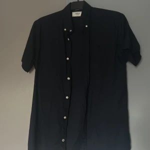 Mörkblå kortärmad skjorta från Produkt - Säljer en stilren mörkblå skjorta från Produkt med korta ärmar och klassisk krage. Skjortan har vita knappar framtill och en bröstficka. Perfekt för dig som gillar enkel och clean stil.