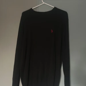 Svart stickad tröja från US polo - Svart stickad tröja från U.S Polo med klassisk rund halsringning och röd broderad logga på bröstet. Långärmad modell i mjukt material som passar perfekt till lager-på-lager. Enkel och stilren design.