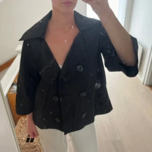 Svart skepparkavaj med stora knappar - Stilren svart skepparkavaj med bred krage och dubbelknäppning framtill. Jackan har trekvartsärmar och en loose passform som ger en avslappnad men ändå snygg look. Perfekt för dig som vill ha en klassisk jacka med modern twist.