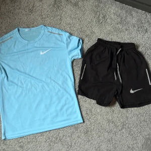Ljusblå Nike set - Nike t-shirt i ljusblått och storlek XS, med klassisk vit swoosh-logga på bröstet och reflekterande detaljer på axlarna. Tillverkad i ett lätt och ventilerande funktionsmaterial, perfekt för träning. Rund halsringning och korta ärmar ger en sportig vibe.