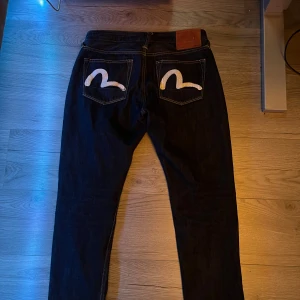 Mörkblå jeans från Evisu, 31x34 - Säljer ett par mörkblå jeans från Evisu med deras ikoniska vita målad logga på bakfickorna. Jeansen har klassisk rak passform, orange kontrastsömmar och är tillverkade i kraftigt denimtyg. Perfekta för dig som vill sticka ut med en unik streetwear-look.