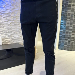 Svarta byxor från Just Junkie M - Svarta byxor från Just Junkie i slim passform med dragkedjedetaljer på sidorna. Byxorna har en modern, avsmalnande siluett och är tillverkade i ett mjukt bomullsmaterial. Perfekta för dig som gillar en stilren look med edge.