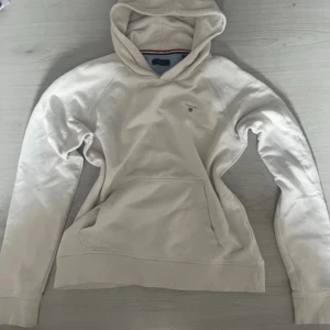 Vit hoodie från GANT med logga - Snygg vit hoodie från GANT med broderad logga på bröstet. Klassisk känguruficka framtill, huva och mjukt bomullsmaterial. Perfekt för en chill och stilren look. Använt 2 ggr 