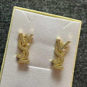 YSL örhängen i guld med strass - Snygga YSL örhängen i guldpläterad metall med glittrande strass. Designen är formad som YSL-loggan och har en lyxig vibe med små stenar som ger extra bling. Perfekt för dig som vill sticka ut med ett statement-smycke från ett ikoniskt modehus.