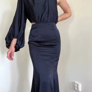 Marinblå långklänning med oneshoulder - Snygg marinblå långklänning i glansigt material med en ärm och oneshoulder-design. Klänningen har ballongärm, markerad midja och cutout i sidan samt öppen rygg. Figurnära passform upptill och utsvängd nertill, perfekt för dig som vill sticka ut. Säljer då storleken inte passar mig längre och jag behöver köpa en ny
