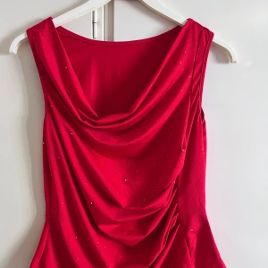 Röd glittrig kortklänning - Snygg röd kortklänning med glittriga detaljer och draperad urringning. Ärmlös modell i mjukt, stretchigt material som sitter bekvämt och ger en festlig look. Perfekt för dig som vill sticka ut med lite extra glitter.