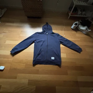 Mörkblå hoodie med dragkedja - Säljer en mörkblå hoodie från Andy med hel dragkedja framtill och huva. Tröjan har långa ärmar, två fickor och en vit patch med logga nertill. Perfekt för chill dagar eller när du vill ha en enkel och stilren look.