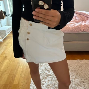 Vit kortkjol med knappar - Snygg vit kortkjol med dekorativa guldknappar framtill och fickor bak. Kjolen har en omlott-look och är perfekt för en trendig och fräsch stil. Passar till både vardagsoutfits och mer uppklädda tillfällen.