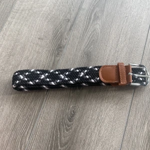 Svart och vit flätat bälte - Snyggt flätat bälte i svart och vitt med bruna skinn-detaljer vid spännet. Bältet har ett klassiskt silverfärgat spänne och passar perfekt till jeans eller chinos för en stilren look.