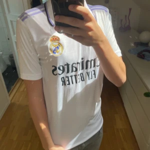 Real Madrid vit matchtröja Adidas - Säljer en officiell Real Madrid fotbollströja från Adidas i vit med lila detaljer på axlarna och svart krage. Tröjan har klubbmärket och sponsortryck på bröstet, samt klassiska Adidas-ränder. Materialet är lätt och andas, perfekt för träning eller match.