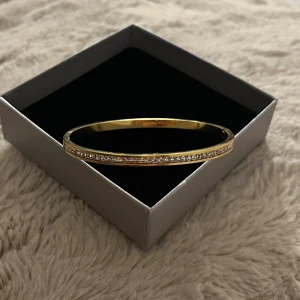 Guldfärgat armband med glittriga stenar - Ett stilrent guldfärgat armband med raka linjer och en rad gnistrande klara stenar längs ovansidan. Armbandet är stelt och har en enkel, elegant design som passar perfekt för att lyfta vilken outfit som helst. Den har inte stenar på baksidan. 