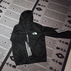 Svart hoodie från The North Face - Svart hoodie från The North Face med vit logga på bröstet och vita ränder på ena ärmen. Tröjan har huva, dragkedja framtill och är långärmad. Perfekt för dig som gillar en sportig och clean stil.