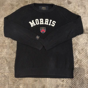 Svart stickad tröja från Morris - Svart stickad tröja från Morris med vit broderad text och ett färgglatt emblem på bröstet. Tröjan har rund halsringning och långa ärmar, perfekt för kyliga dagar. Klassisk och stilren design som funkar till många outfits.